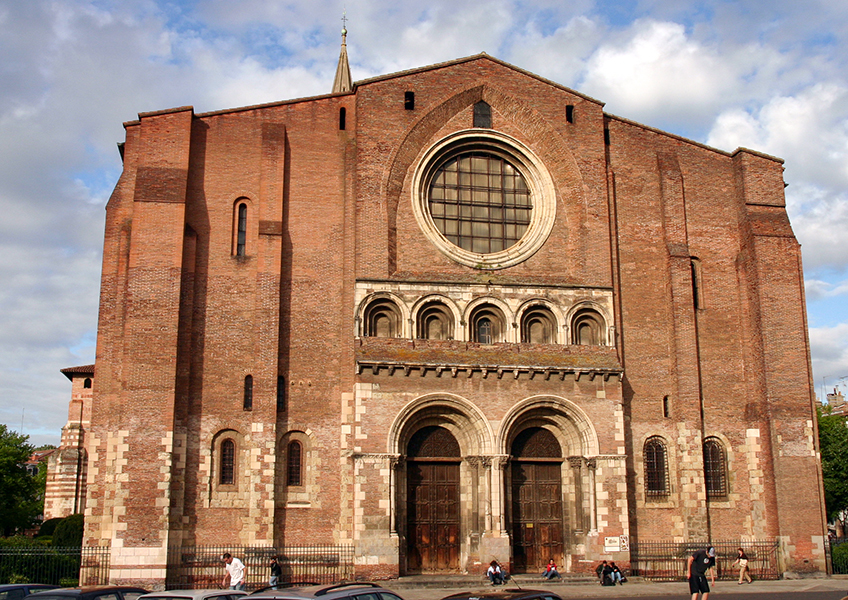 Photograph of a large historic brick church facade featuring a prominent circular stained glass window above three arched doorways. The structure includes vertical buttresses, small rectangular windows, and a few people and parked cars in front, highlighting its scale and architectural details.