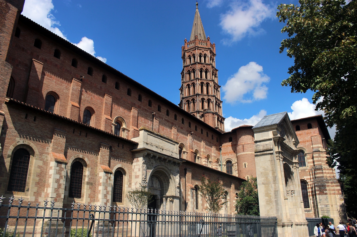 Photograph of a historic church with a tall, ornate bell tower under a blue sky with scattered clouds. The structure features arched windows, detailed stonework, and a wrought iron fence in the foreground, highlighting architectural elements typical of Romanesque style.