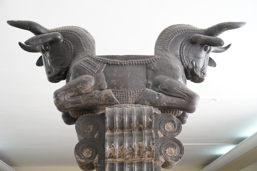 Photograph of a Bull Capital from Persepolis sculpture featuring two symmetrical bull heads facing opposite directions, mounted on a decorated column. The sculpture showcases intricate carvings on the bulls' bodies and the column base, highlighting ancient artistic craftsmanship and cultural symbolism.