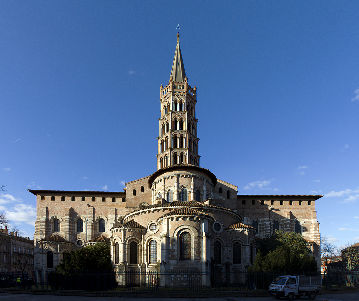 Photograph of a historic Romanesque church with a tall, ornate bell tower under a clear blue sky. The building features rounded arches, detailed stonework, and symmetrical design, with shadows emphasizing architectural depth and a parked vehicle visible in the foreground.