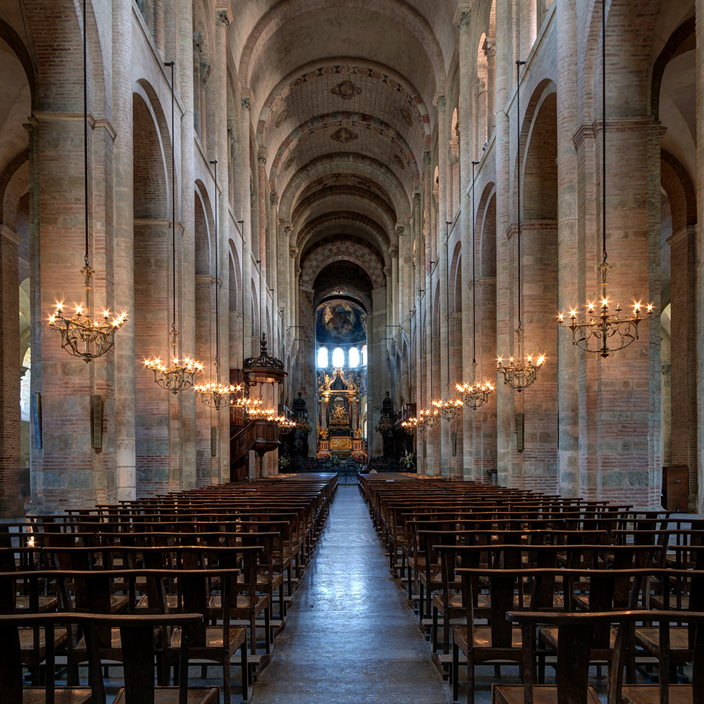 Photograph of a long, vaulted church interior with rows of wooden pews aligned on both sides of a central aisle leading to an ornate altar. Warm lighting from wall-mounted chandeliers highlights stone arches and detailed ceiling fresco in the apse.