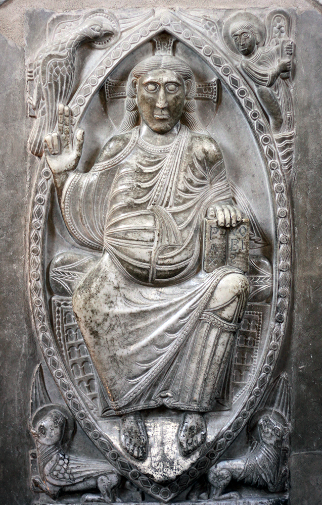 A carved stone relief depicting a seated religious figure within an almond-shaped mandorla, surrounded by symbolic creatures including a lion, an ox, an eagle, and a human figure. The figure holds a book with inscribed letters and raises a hand in blessing, highlighting themes of divinity and authority in Christian iconography.