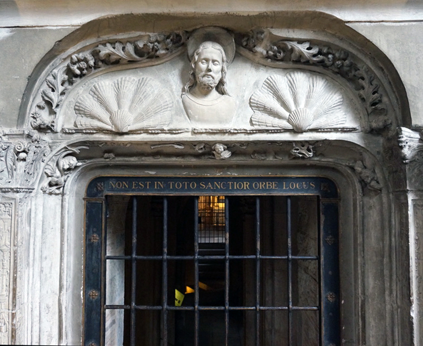 Photograph of an ornate stone doorway featuring a barred gate and Latin inscription above the entrance. Decorative elements include two large carved shells and a central bust sculpture framed by intricate floral motifs.