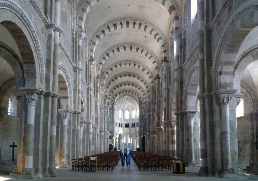 Photograph of a large Romanesque church interior featuring tall stone columns and rounded arches with alternating light and dark stone patterns. Rows of wooden chairs line the central aisle leading to an illuminated altar area, with two people walking toward it, highlighting the building's grand scale and architectural symmetry.