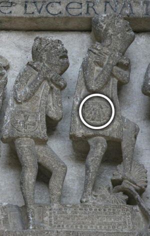 A photograph of a stone relief sculpture depicting two human figures carrying heavy loads on their backs. The sculpture features detailed carvings, including a circular emblem on the load of the right figure and an inscription above the figures.
