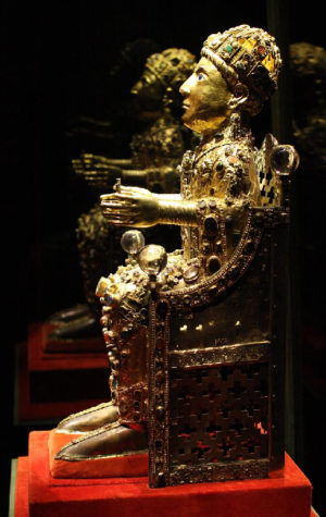 Photograph of an ornate gold and jewel-encrusted statue seated on a detailed throne, displayed on a red pedestal. The statue features intricate craftsmanship with embedded gemstones and a reflective dark background highlighting its elaborate design and regal posture.