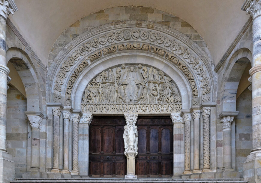 Photograph of a Romanesque church portal featuring intricately carved stone arches and a central wooden door. Detailed sculptures include biblical figures and decorative patterns, with a prominent statue positioned on a column in front of the door.