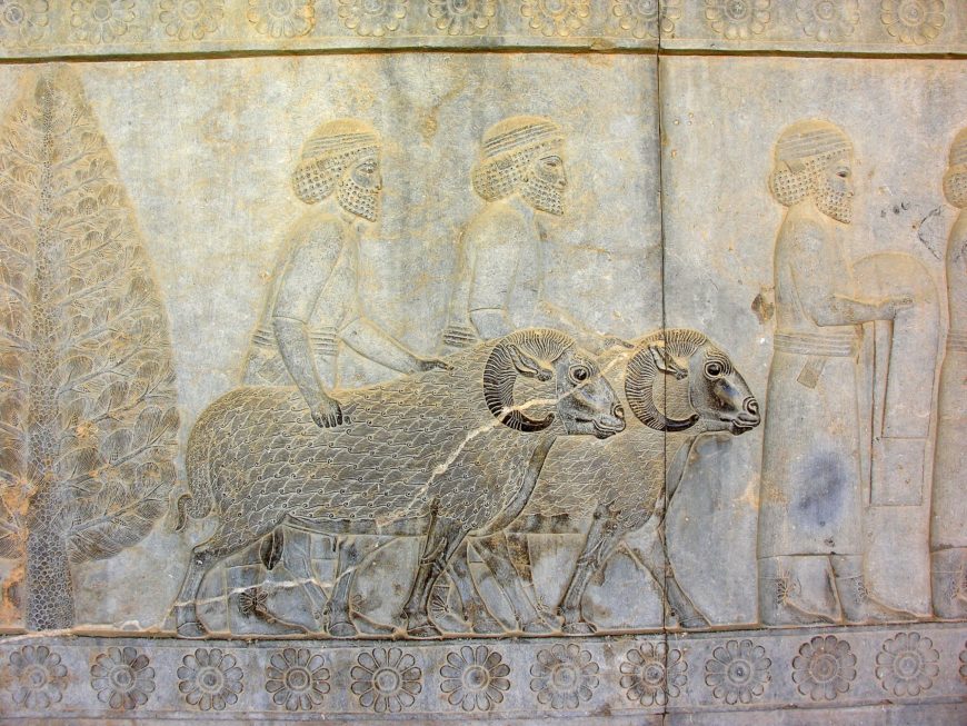 Assyrians with Rams bringing tribute to the Persian king bas-relief carving depicting three men leading two rams, with detailed patterns on animals and figures, set against a floral border and a tree on the left. This ancient stone artwork emphasizes intricate textures on hair, clothing, and animal fur.