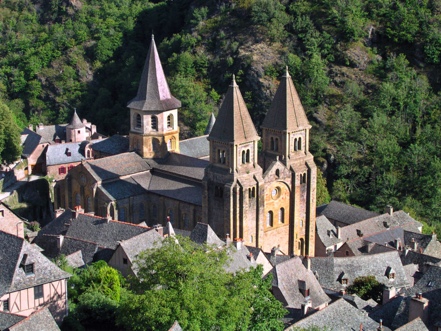 Photograph of a historic stone church with two tall spires and a smaller bell tower, surrounded by traditional slate-roofed buildings nestled in a green, forested hillside. The scene highlights architectural details of the church and the quaint village setting, emphasizing the contrast between man-made structures and natural landscape.