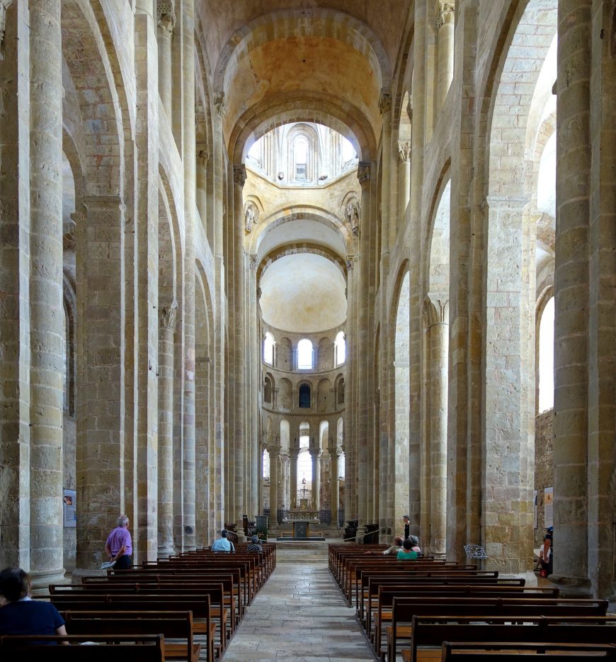 Photograph of a Romanesque church interior featuring tall stone columns, rounded arches, and a central aisle leading to an altar with a crucifix. Natural light filters through arched windows, highlighting the architectural symmetry and spacious nave with wooden pews on both sides.