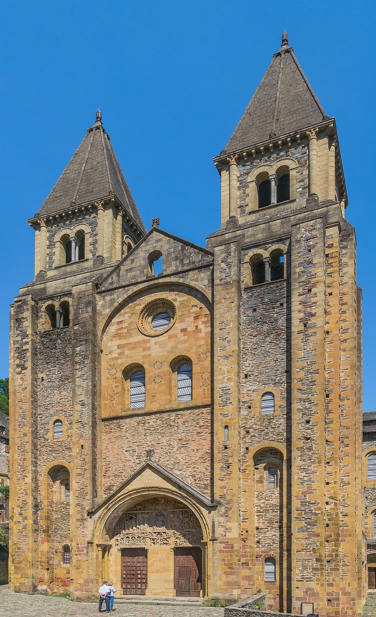 Photograph of a Romanesque church facade featuring two tall, pointed towers with arched windows and a central section with a circular window above three smaller arched windows. The stone structure displays detailed carvings above the entrance and a clear blue sky background.