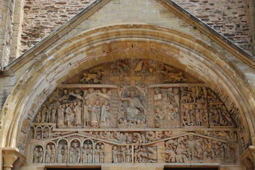 Photograph of a detailed stone tympanum above a church entrance, depicting religious scenes with multiple human figures arranged in horizontal registers. Central figure seated with a cross behind, surrounded by angels, apostles, and various narrative scenes carved in relief within an arched frame of weathered stone.