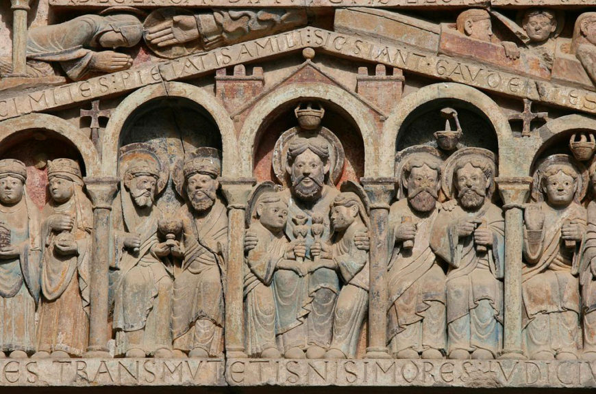 A photograph of a medieval stone relief sculpture depicting a series of religious figures arranged in arched niches with Latin inscriptions above and below. The relief features detailed carvings of robed individuals, some holding objects, with a central figure elevated above others, highlighting its significance in the composition.