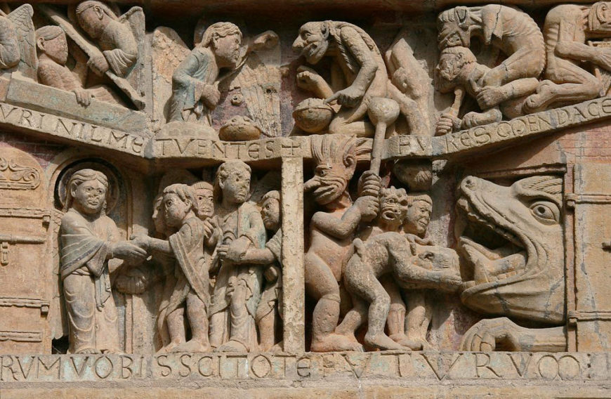 A detailed stone relief sculpture depicting a biblical scene with multiple human and monstrous figures interacting, including a large creature with an open mouth on the right. Latin inscriptions run along top and bottom edges, with intricate carvings showing various postures and expressions, highlighting dramatic narrative elements.