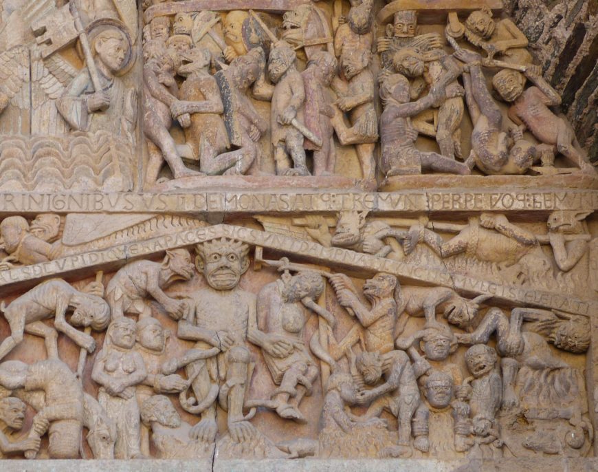 A detailed stone relief sculpture depicting a chaotic scene with numerous human and demonic figures engaged in various actions, representing a religious narrative. The carving features Latin inscriptions, layered composition with a large central demonic figure, and intricate expressions and gestures highlighting themes of torment and struggle.