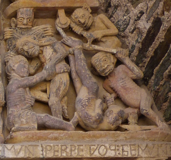 A photograph of a detailed stone relief sculpture depicting five grotesque, humanoid figures in a chaotic scene, possibly representing a religious narrative. The relief features intricate textures, exaggerated facial expressions, and a carved inscription along the bottom edge.