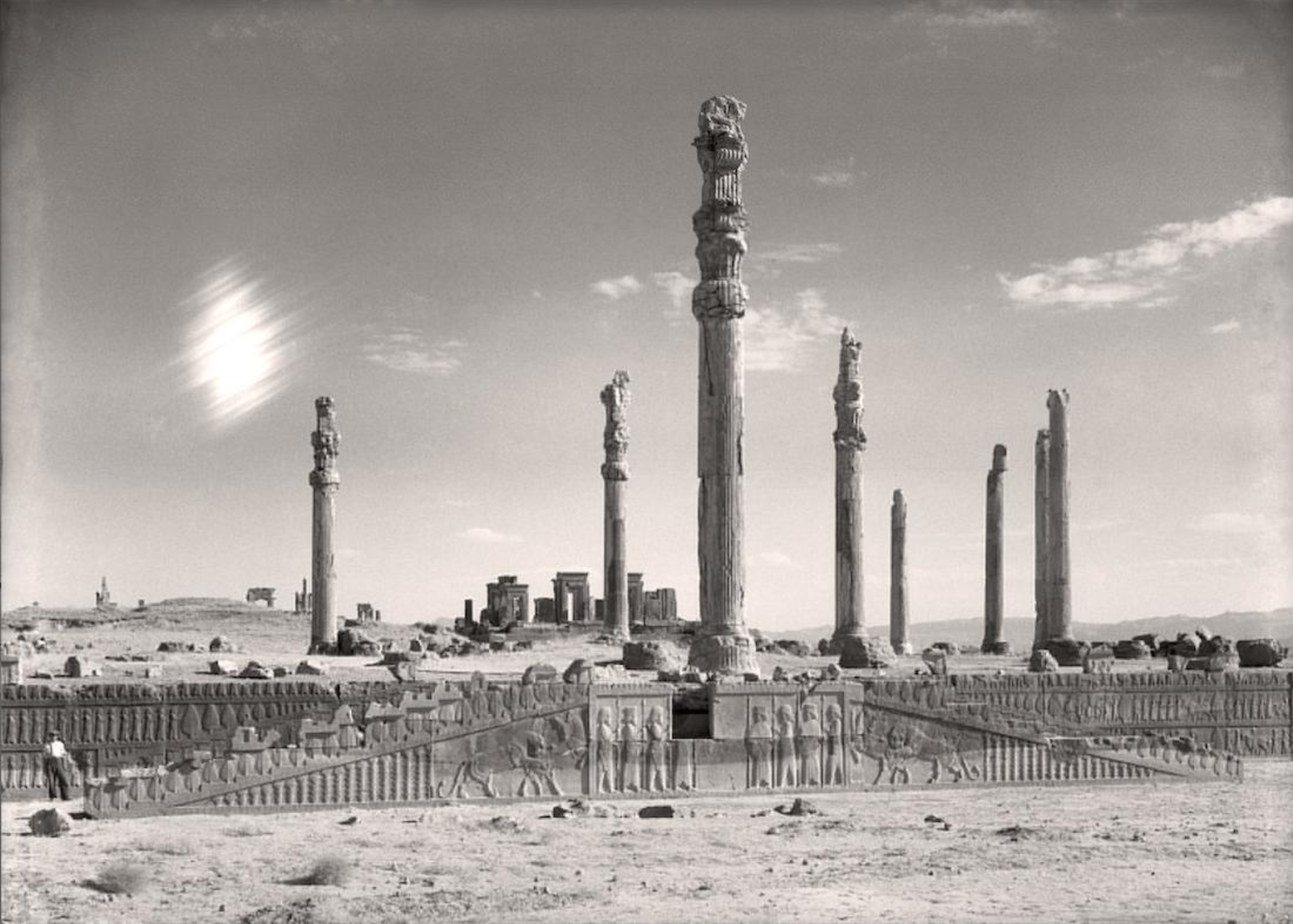 Black and white photograph of View of the eastern stairway and columns of the Apādana, Persepolis featuring tall, intricately carved stone columns arranged in a row with detailed bas-reliefs on surrounding walls. The scene captures a wide, open archaeological site under a partly cloudy sky.