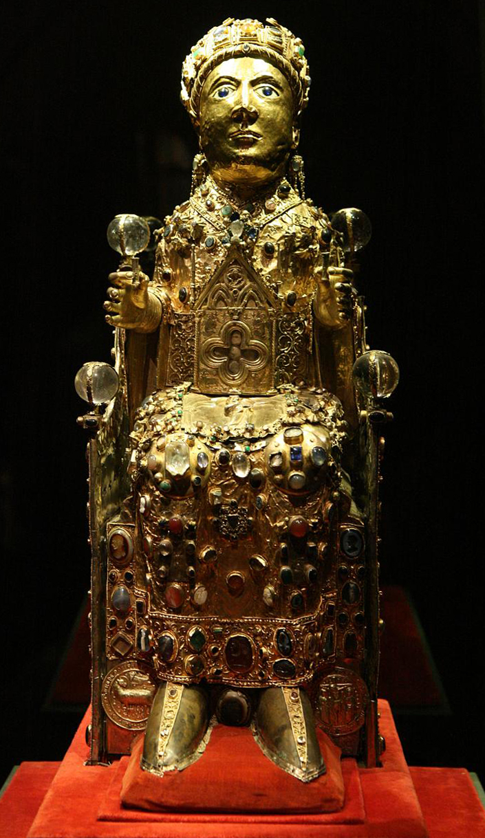 Photograph of an ornate medieval reliquary statue depicting a seated figure adorned with gold, jewels, and intricate metalwork. The figure holds two orbs and wears a crown, with detailed embellishments covering the robe and throne.