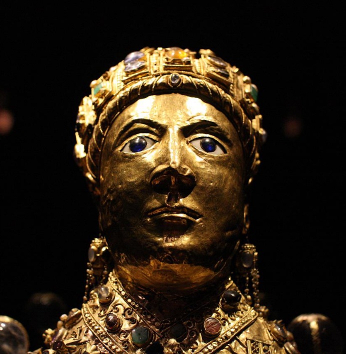 Photograph of an ornate golden bust adorned with various colorful gemstones and intricate metalwork, set against a dark background. The bust features detailed craftsmanship with embedded jewels and decorative patterns.
