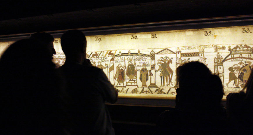 Photograph of people viewing a section of the illuminated Bayeux Tapestry displayed in a museum setting. The tapestry depicts medieval scenes with labeled numbers, featuring figures in period clothing and architectural elements, illustrating historical events from the Norman Conquest.