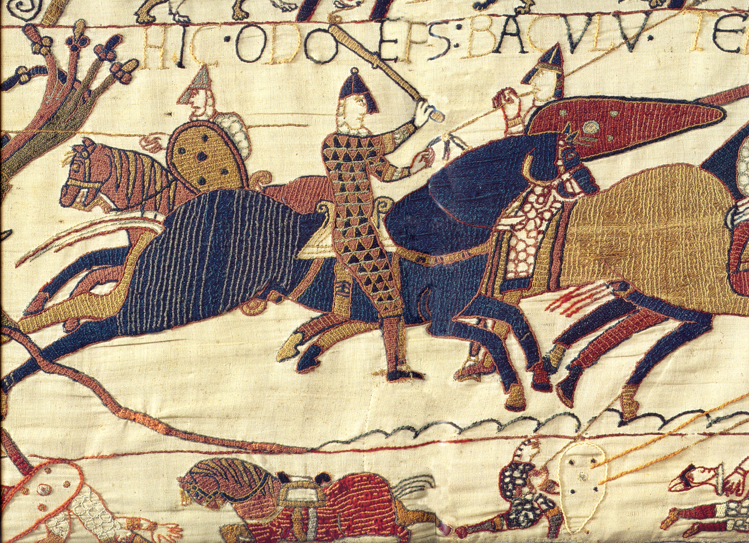A photograph of a medieval embroidered tapestry depicting a battle scene with mounted soldiers wielding swords and shields. The tapestry features detailed patterns on horses and armor, with Latin inscriptions above and a muted color palette of blues, reds, and browns.