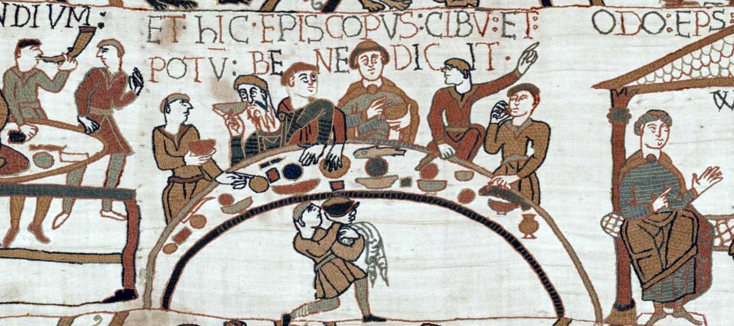 A medieval embroidered tapestry scene depicting a banquet with several seated figures eating and drinking at a long, curved table, accompanied by a servant carrying a jug underneath. The scene includes Latin text above the figures and features detailed clothing and tableware in muted colors on a beige fabric background.
