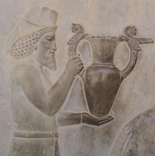 An Armenian tribute bearer carrying a metal vessel with Homa (griffin) handles bas-relief sculpture depicting a bearded man wearing a hat, holding a large, ornate vessel with animal-shaped handles.