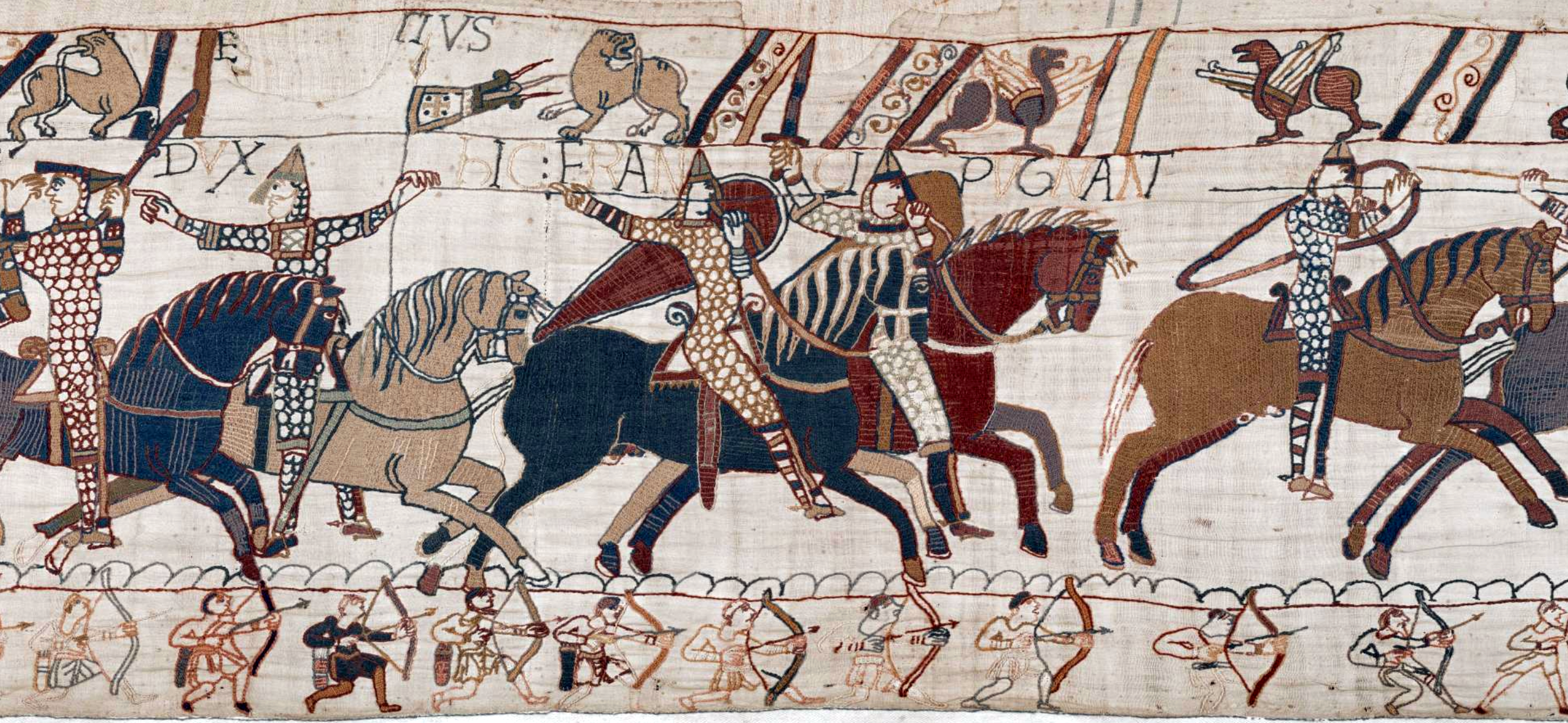 A medieval embroidered tapestry depicting a mounted battle scene with knights in armor wielding lances and shields, accompanied by foot soldiers below. The artwork features detailed patterns on horses and armor, with Latin inscriptions and symbolic figures above the riders.