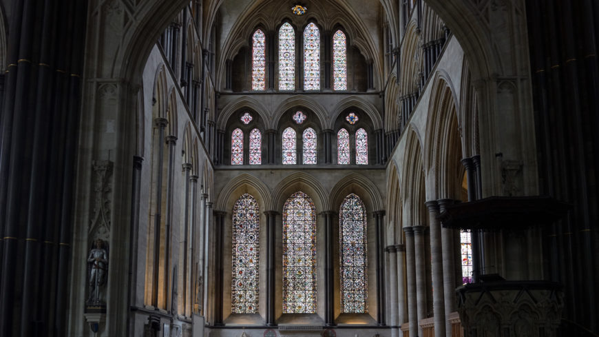 Photograph of a Gothic cathedral interior showcasing tall, pointed arches and multiple stained glass windows arranged in two tiers. The image highlights intricate stone columns, detailed tracery in windows, and soft natural light filtering through colorful glass panels.