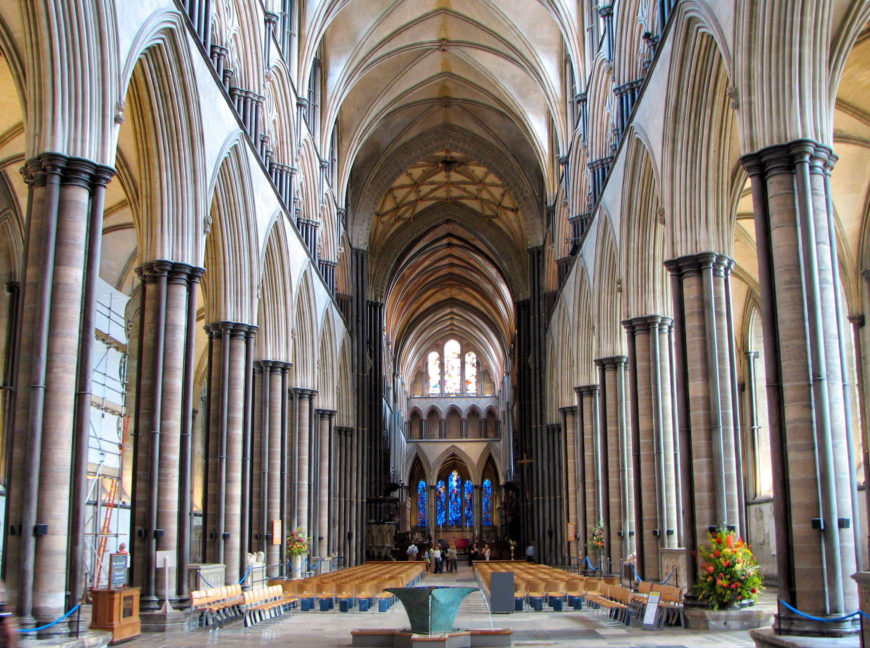 Photograph of a cathedral interior showcasing Gothic architecture with tall, pointed arches, ribbed vaults, and clustered columns. Symmetrical rows of wooden pews lead to a stained glass window and altar, with floral arrangements on either side.