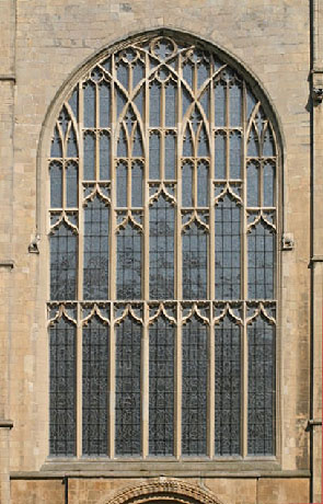 Photograph of a large Gothic-style stained glass window set in a stone wall, featuring intricate vertical and pointed arch tracery. The window is divided into multiple tall, narrow panels with detailed stone mullions and decorative elements near the top.