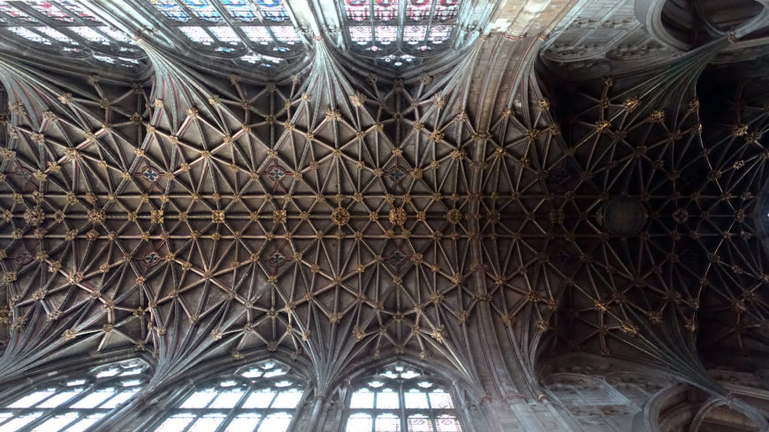 Photograph showing a detailed view of a Gothic vaulted ceiling with intricate ribbed patterns and stained glass windows. The ceiling features complex stonework with intersecting ribs forming star-like shapes.