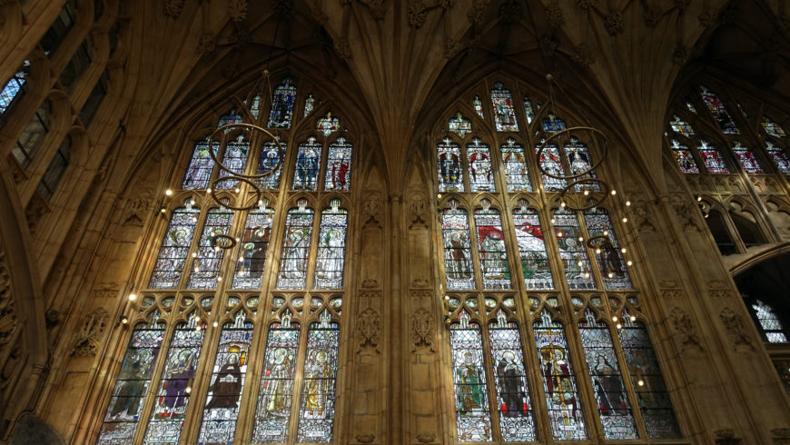 Photograph of large stained glass windows inside a Gothic-style cathedral, showcasing intricate designs and vibrant colors. The windows feature multiple panels with religious figures and scenes, framed by detailed stone arches and ribbed vaulting above.