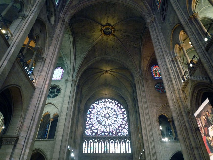 Photograph of a Gothic cathedral interior showcasing tall stone arches, ribbed vaulted ceilings, and a large circular stained glass rose window with intricate colorful patterns. Light filters through smaller stained glass windows on either side, highlighting architectural details and creating a serene atmosphere.