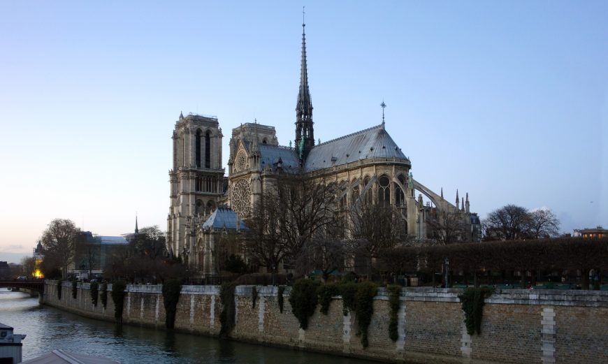 Photograph of Notre-Dame Cathedral in Paris taken from across the Seine River during early evening, showcasing its iconic Gothic architecture with twin towers and central spire. Surrounding trees and stone embankments frame the historic landmark under a clear sky.