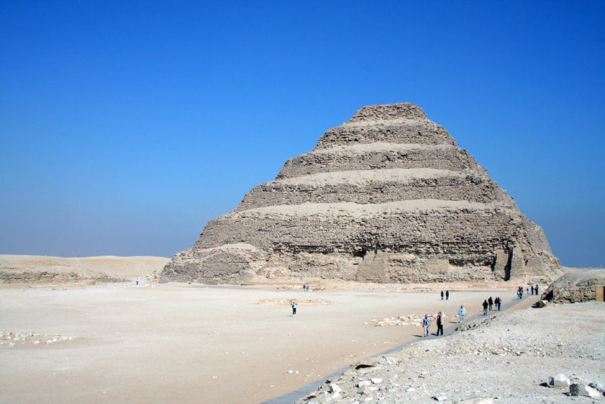 Photograph of the Step Pyramid of Djoser in Egypt, showcasing its distinct layered structure and surrounding desert landscape under a clear blue sky. Several small groups of people near the pyramid provide scale, highlighting its massive size and historical significance as one of the earliest large stone structures.