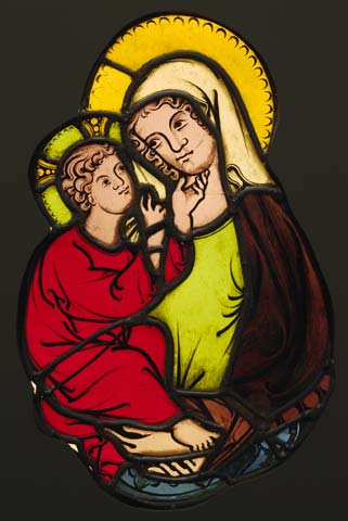 Stained glass artwork depicting a religious scene with two haloed figures, one in red robes and the other in green and brown garments, suggesting a depiction of Madonna and Child. The piece features intricate black outlines and vibrant colors, emphasizing the tender interaction between the figures.