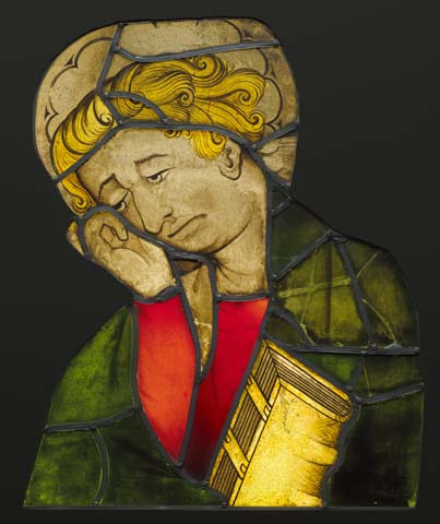 A stained glass artwork depicting a figure with golden hair, wearing a green robe and red garment, holding a yellow book. The design features dark leading lines separating colored glass pieces, highlighting the figure's contemplative pose and detailed hair texture.
