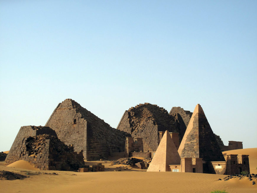 Photograph of ancient Nubian pyramids located in a desert landscape under clear blue sky. The scene features several stone pyramids in various states of preservation, with some showing significant erosion and others more intact, highlighting historical architectural styles.