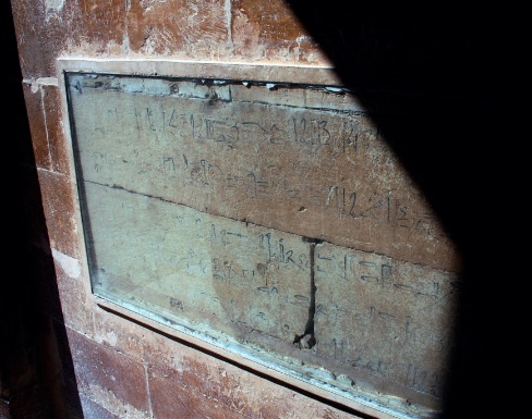 Photograph of an ancient graffiti on stone embedded in a wall, featuring faded inscriptions or symbols. The stone is framed with a protective glass cover, with visible cracks and weathering. .