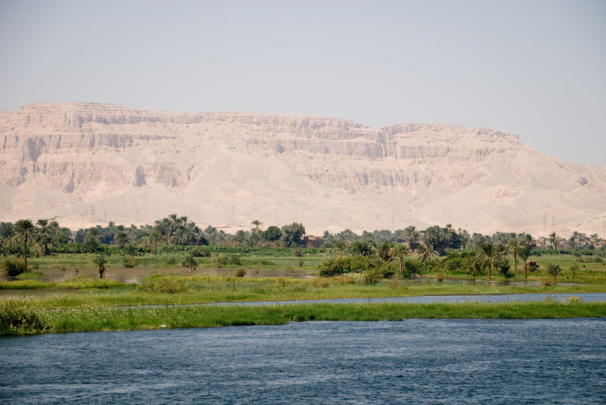 Photograph of the Nile river with lush green vegetation and palm trees along its banks, set against a backdrop of large, beige rocky cliffs under a clear sky. The scene highlights contrast between fertile riverbanks and arid desert landscape looking towards the Valley of Kings.