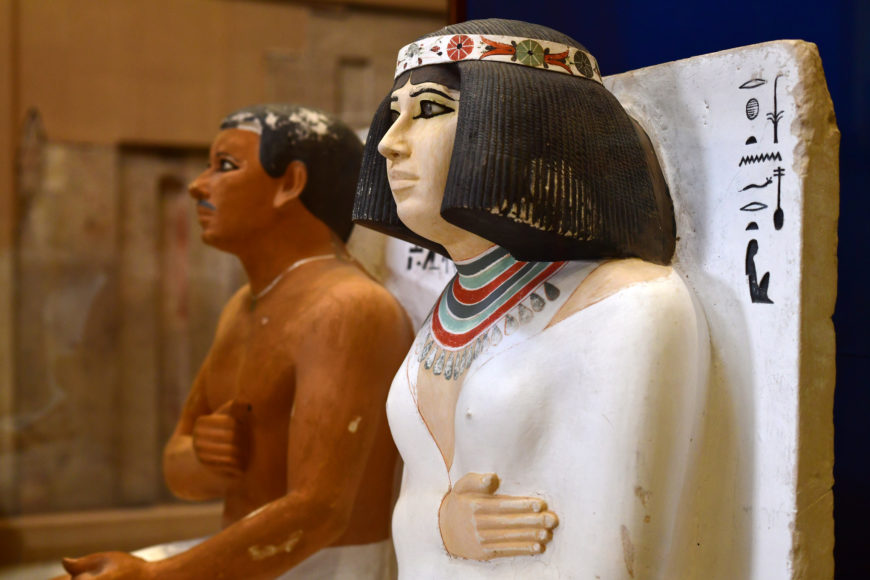 Photograph of two ancient Egyptian statues displayed side by side, depicting male and female figures with detailed attire and painted features. The statues showcase traditional Egyptian artistic styles, including hieroglyphics on the white statue's back panel and intricate jewelry painted around the neck.
