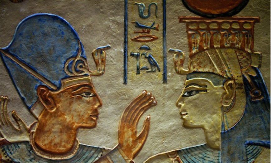 A photograph of an ancient Egyptian wall relief depicting two figures facing each other, one wearing a blue headdress and the other adorned with a crown featuring a sun disk and horns. The relief includes hieroglyphic inscriptions between the figures and showcases detailed carvings with colors such as blue, gold, and brown.