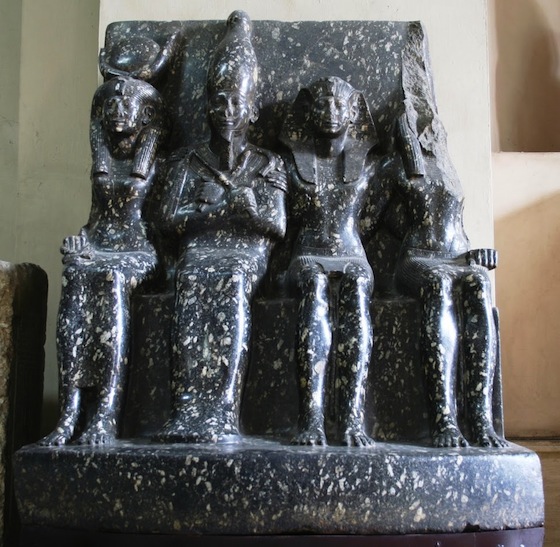 Photograph of an ancient Egyptian stone sculpture depicting four seated royal figures carved from dark speckled stone. The central figure wears a tall headdress and holds crossed arms, flanked by three figures with distinct postures and headgear, all seated on a block-like throne.