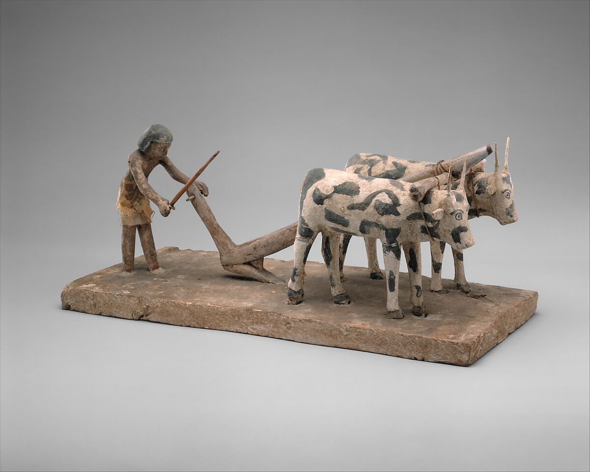 A photograph of an ancient wooden sculpture depicting a farmer plowing a field with two oxen. The sculpture features painted details on oxen with black spots and a farmer holding a plow, mounted on a rectangular base.