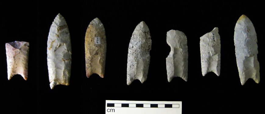 Photograph showing seven ancient stone projectile points arranged in a row on a black background. A scale bar with centimeter markings below the points indicates their varying sizes and shapes, highlighting differences in craftsmanship and wear.
