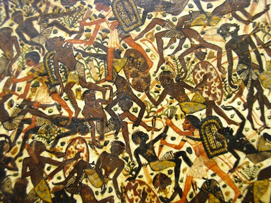 A detailed photograph of an ancient Egyptian wall painting depicting a chaotic battle scene with numerous warriors engaged in combat. The artwork features dark-skinned figures with yellow and brown accents, holding shields and weapons, arranged densely across a beige background with intricate patterns.