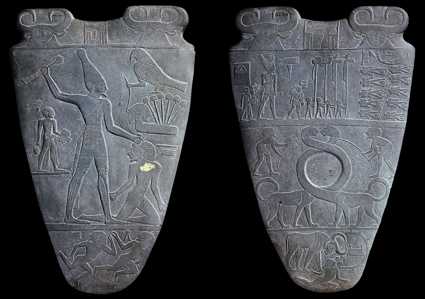 Photograph of an ancient Egyptian ceremonial Palette of King Narmer carved from dark stone, featuring detailed reliefs on both sides. One side depicts a large figure wearing a tall crown holding a mace over a kneeling captive, while the other side shows rows of prisoners, animals, and hieroglyphic symbols arranged in horizontal registers.
