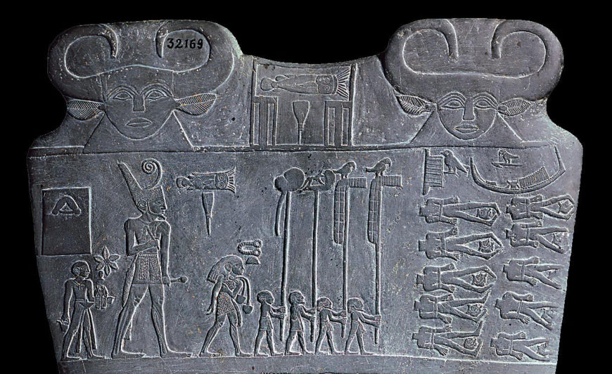 Photograph of detail of Palette of King Narmer stone relief depicting Egyptian figures, including a large central royal figure with a tall headdress, smaller human figures, and hieroglyphic symbols. Relief features two large cow heads at the top corners, detailed carvings of people in procession, and symbolic elements arranged in horizontal and vertical sections.