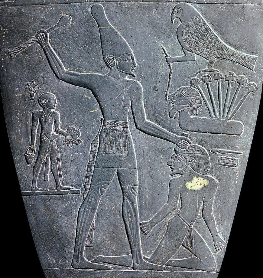 Bas-relief carving Palette of King Narmer depicting an ancient Egyptian scene with a tall figure wearing a tall crown holding a club, standing over a kneeling figure. To the right, a falcon perched on a stylized plant and a smaller figure holding objects are also visible, highlighting themes of power and divinity.